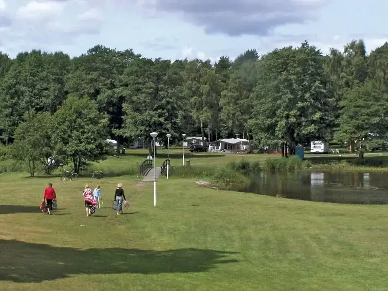Campinggäster på gräset vid dammen med husvagnar i bakgrunden
